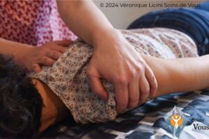 Gros plan sur les mains de Véronique Licini — soin en Ortho-Bionomy® sur l’épaule d’une femme allongée toute habillée